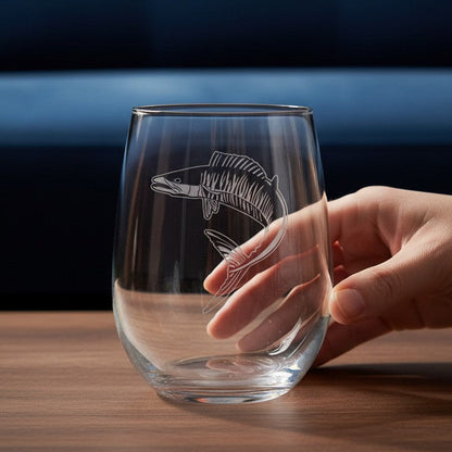 A clear stemless wine glass with an etched ono fish design, held by a hand on a wooden table with a dark blue tufted sofa in the background.