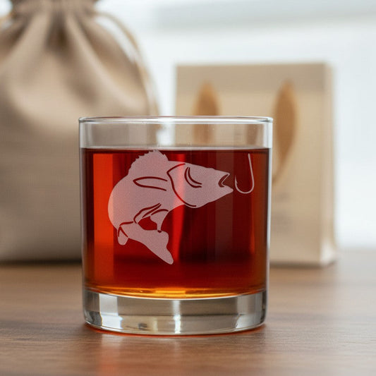 A clear lowball-style tumbler glass filled with amber liquid, featuring a frosted fish-and-hook engraving on the front, set on a wooden surface with a beige drawstring pouch and blurred box in the background.