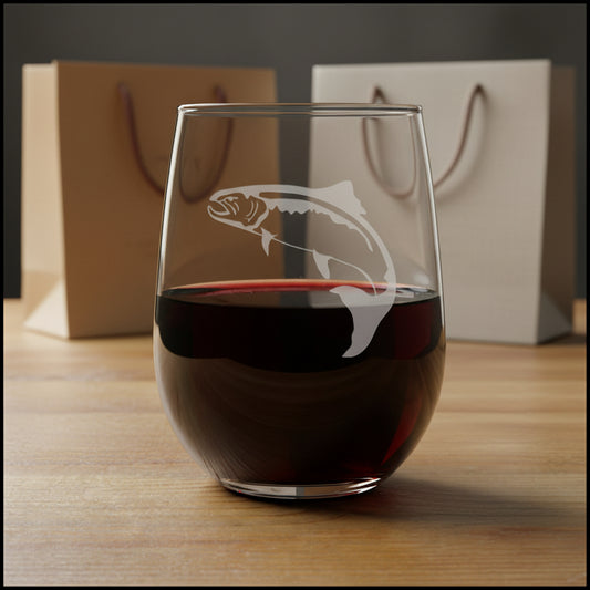 A clear stemless wine glass with an etched salmon design, shown on a wooden surface against a  background of gift bags.