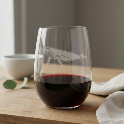 A clear stemless wine glass with an etched whale design, partially filled with red wine, set on a wooden table with a neutral background.