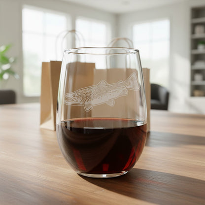 A clear stemless wine glass with an etched trout design, partially filled with red wine, sitting on a wooden table in a bright, modern room with blurred paper bags in the background.