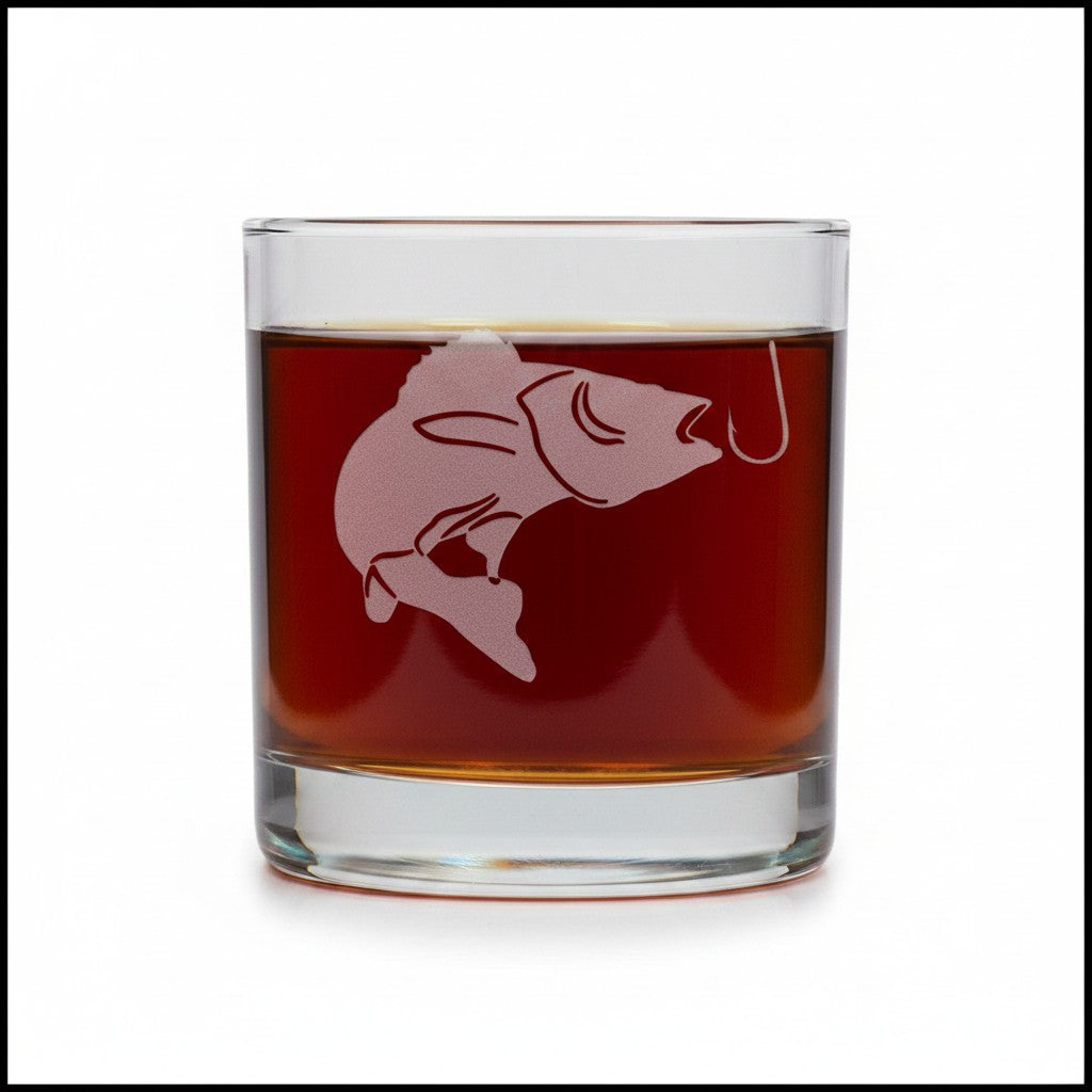 A clear rocks glass filled with amber-brown liquid, featuring a frosted pink fish and hook design on the front, set against a plain white background.