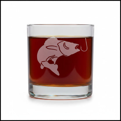A clear rocks glass filled with amber-brown liquid, featuring a frosted pink fish and hook design on the front, set against a plain white background.