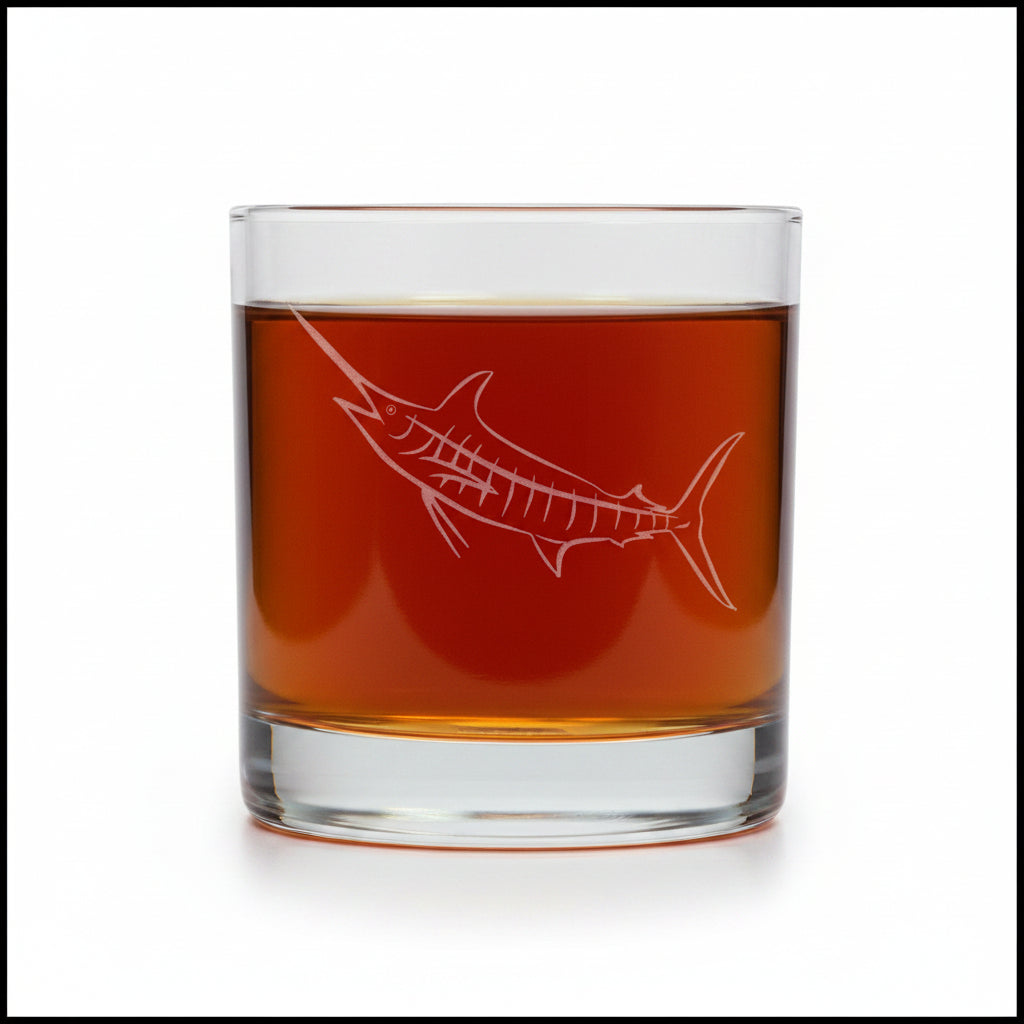 A clear rocks glass with an etched marlin design, filled with a dark red drink, set against a textured gray background.