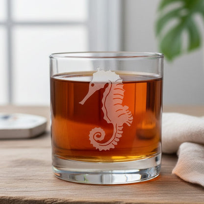 A clear glass rocks tumbler with a etched seahorse etched, filled with amber-colored liquid, set on a wooden surface with a blurred background featuring a plant and window.