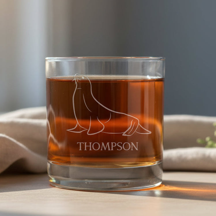 A clear rocks glass tumbler with a white etched seal outline and the name 'THOMPSON' on the front, filled with a dark red liquid.