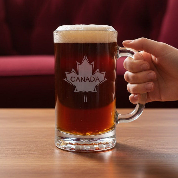A hand holding a clear glass mug filled with amber beer and topped with white foam, featuring a white Canada maple leaf logo on the front, set on a wooden table with a blurred red sofa in the background.