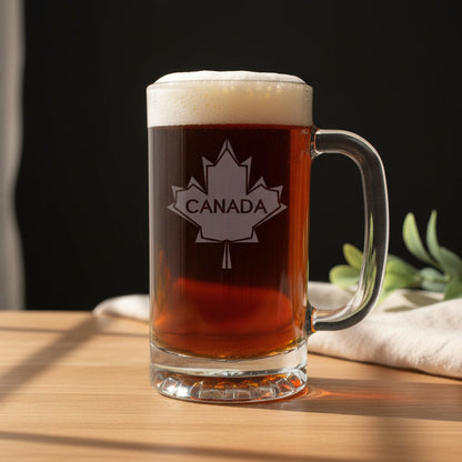 A clear glass beer mug with a handle, filled with dark amber beer and topped with white foam, standing on a light wood surface against a black background. The mug is etched with a white maple leaf containing the word CANADA.