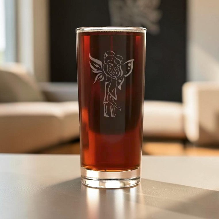 A tall, clear drinking glass filled with a dark reddish-brown liquid, featuring an etched fairy design on the front, set on a reflective tabletop with a blurred living room background.