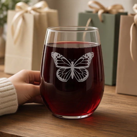 A stemless wine glass filled with red wine, featuring an engraved butterfly design on the front, held by a hand on a wooden table with gift bags in the background.