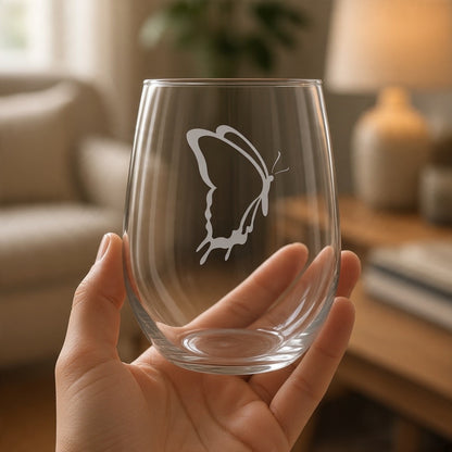 A clear stemless wine glass with an etched butterfly design, held by a person's hand on a wooden table with a living room in the background.