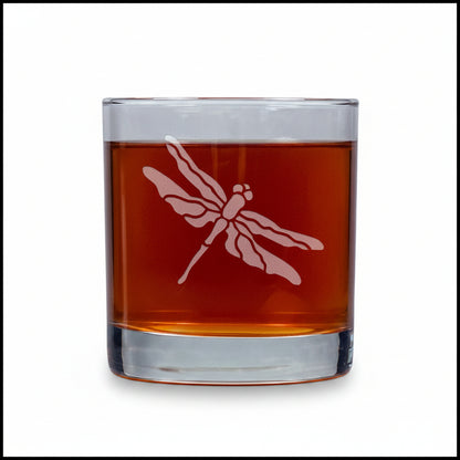 A clear rocks glass with a dragonfly etched design, filled with an amber-brown liquid, set against a white background.