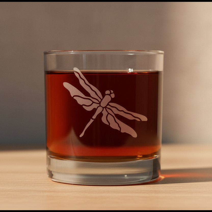A clear rocks glass with a dragonfly etched design, filled with an amber-brown liquid.
