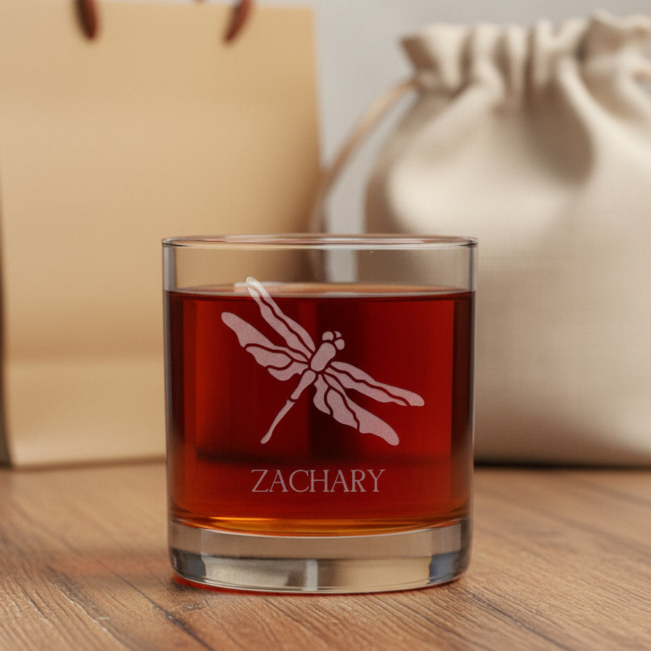 A clear rocks glass filled with amber-red liquid, featuring an etched dragonfly design and the name 'ZACHARY' on the front.