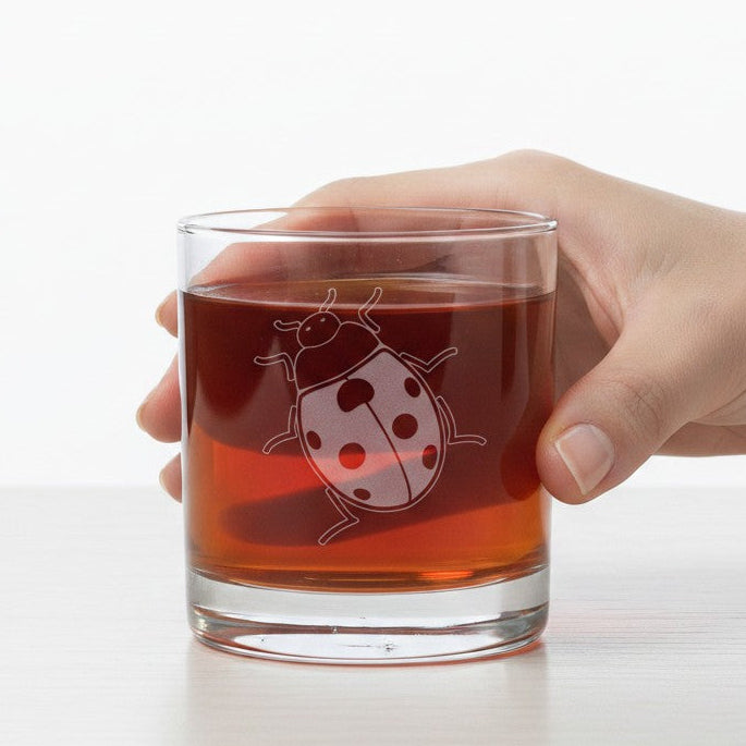 A hand holding a clear lowball glass with an etched  ladybug design, filled with an amber-brown liquid, on a white surface against a white background.