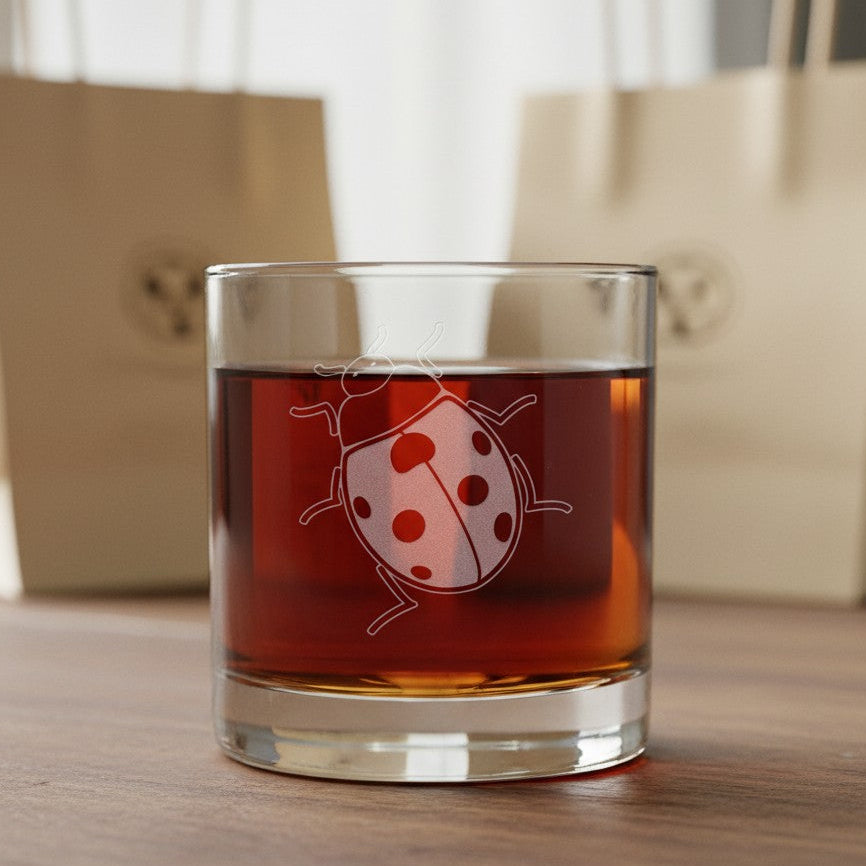A clear rocks glass with a white etched ladybug design, filled with an amber-brown liquid, set on a wooden surface with blurred beige shopping bags in the background.