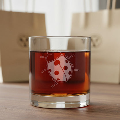 A clear rocks glass with a white etched ladybug design, filled with an amber-brown liquid, set on a wooden surface with blurred beige shopping bags in the background.