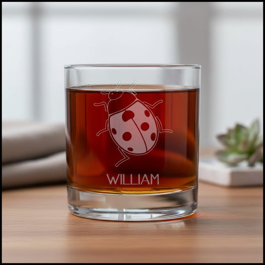 A clear rocks glass filled with amber liquid, featuring an etched ladybug design and the name WILLIAM, set on a wooden tabletop with a softly blurred indoor background.