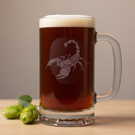 A clear glass beer mug filled with dark amber beer, topped with white foam, featuring an etched scorpion design on the front. The mug sits on a light wood surface against a neutral background.