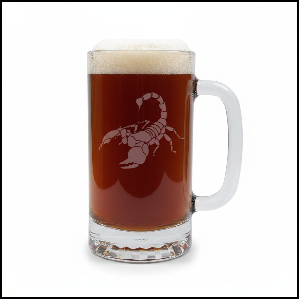 A clear glass beer mug filled with dark amber beer, topped with white foam, featuring an etched scorpion graphic on the front. The mug sits on a light wood surface against a solid white background.