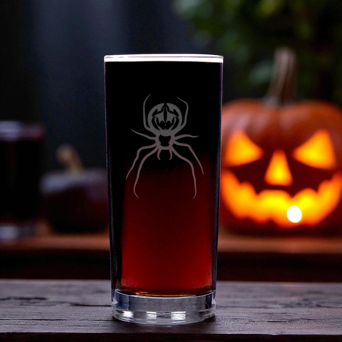 A tall,  cooler glass with an etched spider design, filled with a dark red beverage, set on a wooden surface with a glowing jack-o'-lantern in the background.