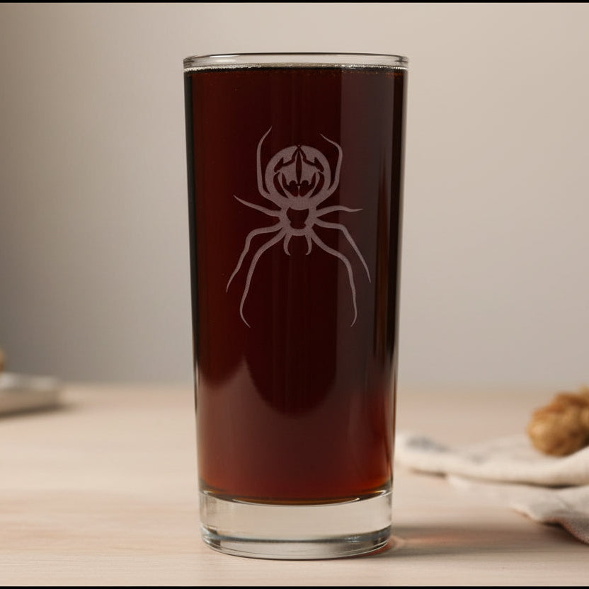 A cooler glass filled with dark beer, featuring a etched spider design on the front, set against a neutral background.