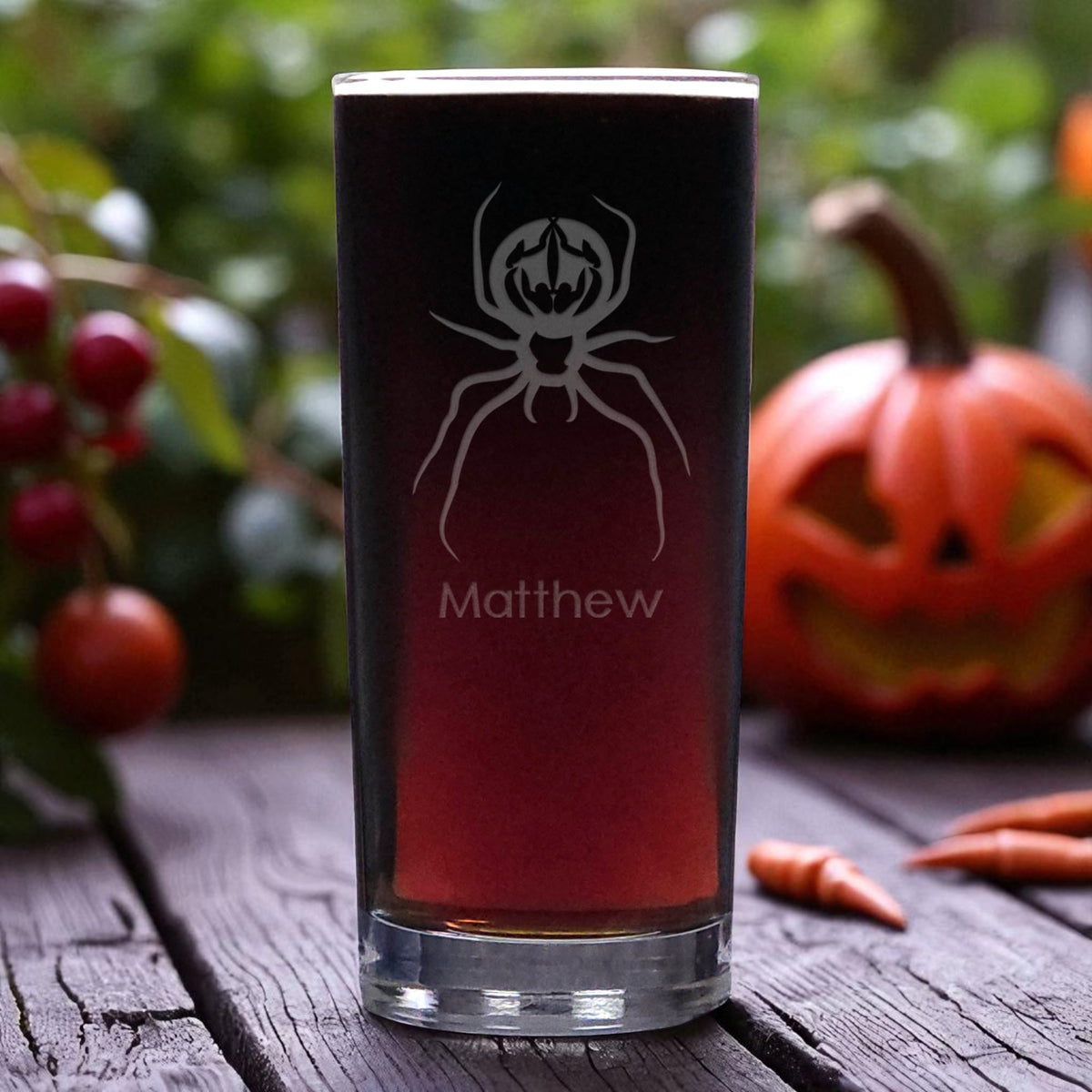 A clear cooler glass  filled with a dark red drink, featuring an etched spider graphic and the name 'Matthew', set on a rustic wooden table with a carved pumpkin and berries in the background.