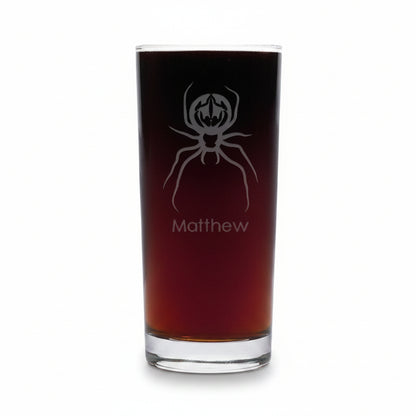 A cooler glass filled with dark red beverage, featuring an etched spider graphic and the name 'Matthew', set against a white background.