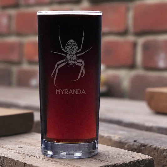 Pint glass with spider design and 'MYRANDA' text on a wooden surface with brick background