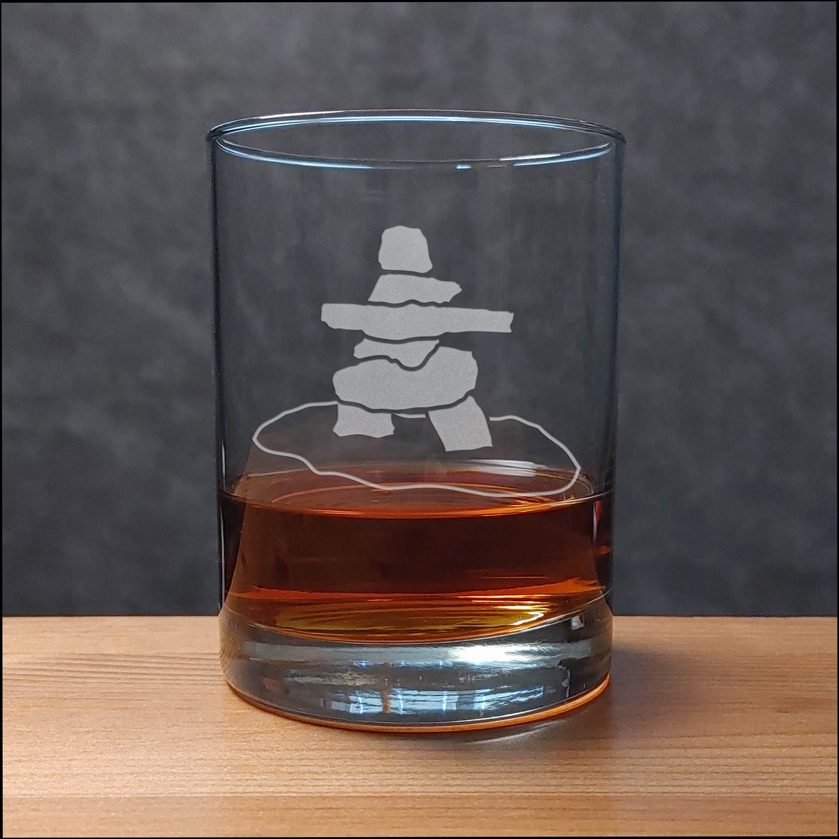 A clear rocks glass with an etched inukshuk (stacked stone) design, partially filled with amber whiskey, sitting on a light wood surface against a dark gray textured background.