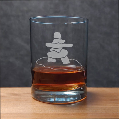 A clear rocks glass with an etched inukshuk (stacked stone) design, partially filled with amber whiskey, sitting on a light wood surface against a dark gray textured background.