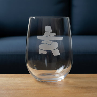 A clear stemless wine glass with a gray etched Inuksuk design, shown on a wooden surface against a blue couch background.