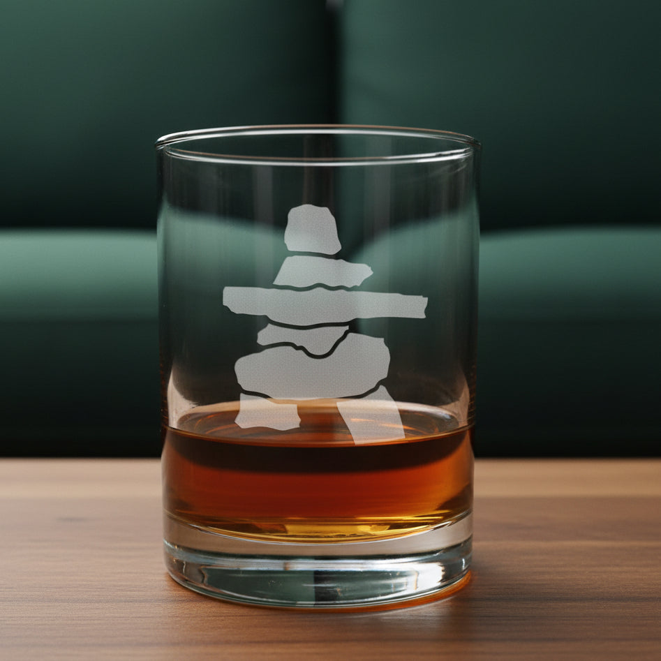 A clear glass rocks tumbler with an etched Inuksuk design, partially filled with amber whiskey, resting on a light wood surface