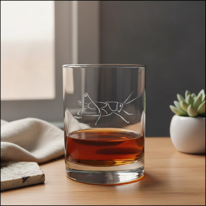 A clear rocks glass with an etched cricket design, partially filled with amber whiskey, set on a wooden table in a softly lit interior with a small potted succulent in the background.