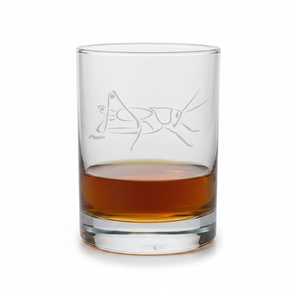 A clear glass rocks tumbler with an etched grasshopper design, partially filled with an amber-brown liquid, shown on a clean white background.
