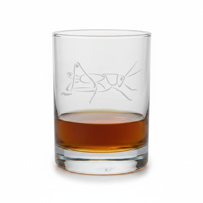A clear glass rocks tumbler with an etched grasshopper design, partially filled with an amber-brown liquid, shown on a clean white background.