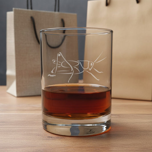 A clear glass rocks tumbler with an etched grasshopper design, filled with amber whiskey, set on a wooden table with kraft paper gift bags in the background.