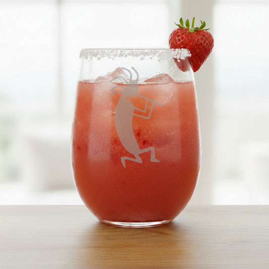 A clear stemless wine glass filled with a cocktail with an etched Kokopelli dancer design, resting on a wooden surface 