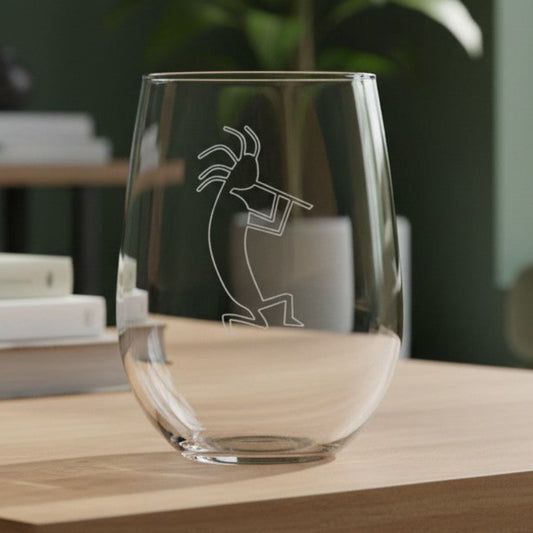 A clear stemless wine glass with an etched Kokopelli figure design, sitting on a light wood table in a modern living room setting.
