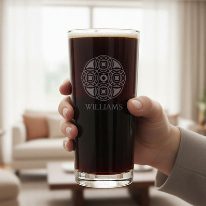 A hand holds a clear cooler glass filled with dark beer, featuring a white mandala circular design and the name WILLIAMS etched on the front, set against a softly blurred living room background.