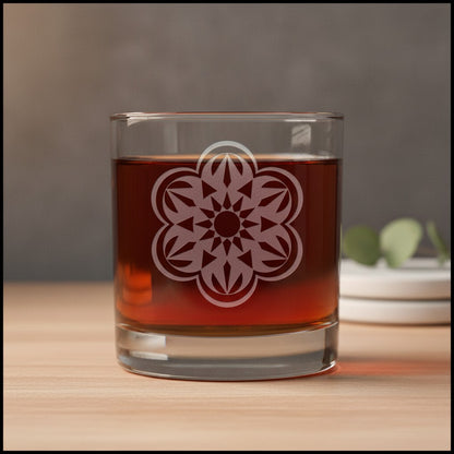 A clear glass rocks tumbler with an etched floral mandala design, filled with an amber-brown liquid, set on a light wood surface against a blurred gray background.