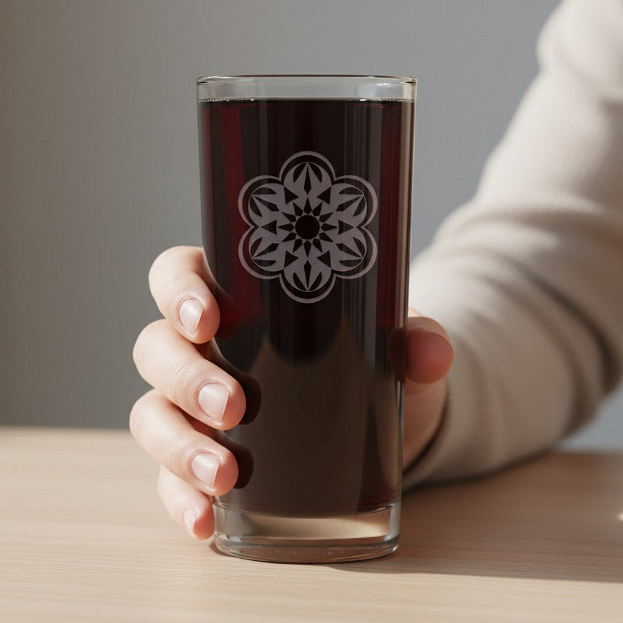 A hand holding a clear glass tumbler with a white floral mandala design, filled with a dark brown beverage, set against a soft-focus indoor background.