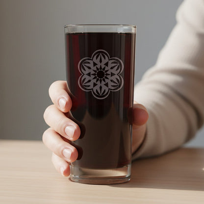 A hand holding a clear glass tumbler with a white floral mandala design, filled with a dark brown beverage, set against a soft-focus indoor background.
