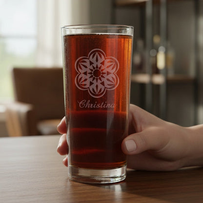 A hand holds a clear cooler glass filled with amber-brown liquid on a wooden table. The glass has a etched floral style mandala design and the name 'Christina' printed below it.