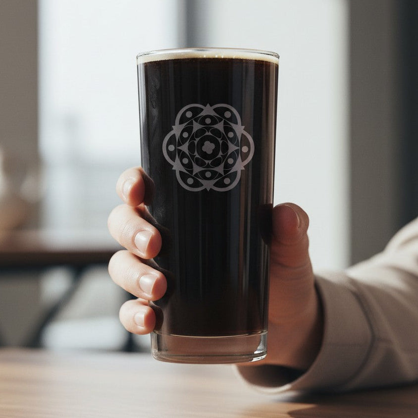 A hand holding a tall clear glass tumbler filled with dark liquid, featuring an etched geometric mandaladesign - set against a softly blurred indoor background.