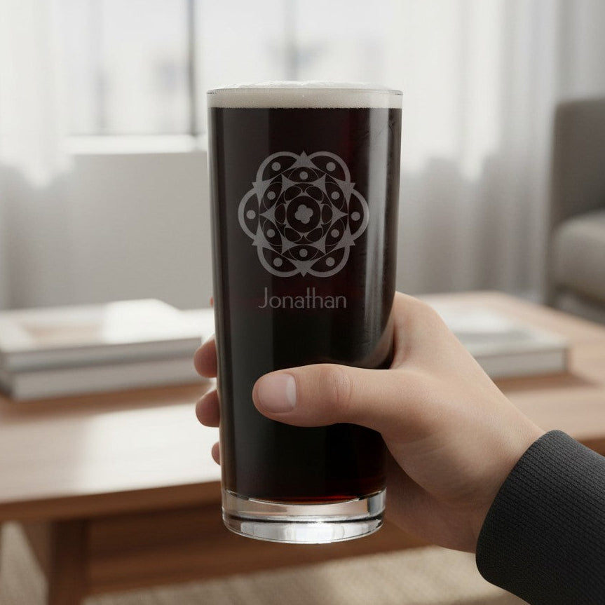 A hand holds a clear glass pint tumbler filled with a dark drink in a softly lit living room. The glass is etched with a circular geometric symbol and the name 'Jonathan'.