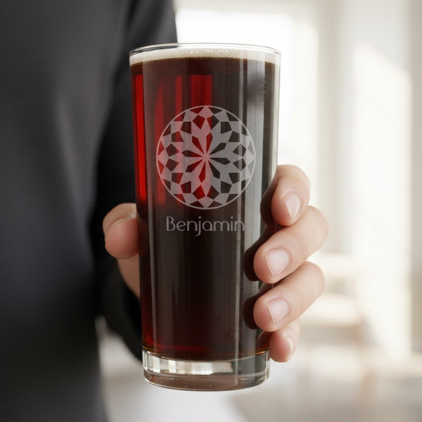 A hand holding a clear glass tumbler filled with dark beer, featuring a red and gray geometric circular logo and the name 'Benjamin' printed below, against a softly blurred indoor background.