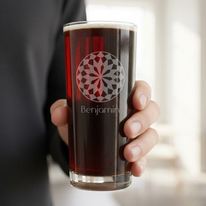 A hand holding a clear glass tumbler filled with dark beer, featuring a red and gray geometric circular logo and the name 'Benjamin' printed below, against a softly blurred indoor background.