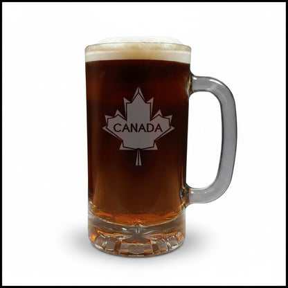 A clear glass beer mug filled with dark amber beer, topped with white foam, featuring a white maple leaf design with the word CANADA on the front, set against a plain white background.