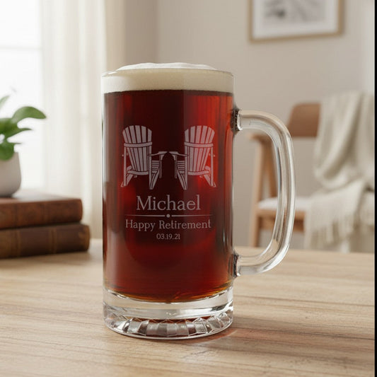 A clear glass beer mug filled with dark red beer and topped with white foam, standing on a wooden table in a softly lit room. The mug is personalized with an etched design of two Adirondack chairs and the name Michael with 'Happy Retirement' and a date.
